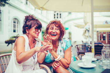 Happy female senior friends laughing and pointing fingers to someone while drinking coffee