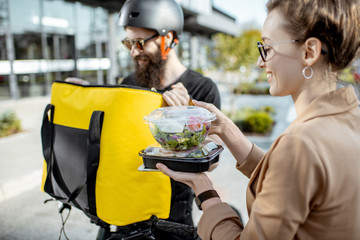 Courier delivering fresh lunches to a young business woman on a bicycle with thermal backpack. Takeaway restaurant food delivery concept