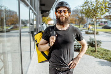 Portrait of a young bearded courier in protective helmet standing with yellow thermal bag for food delivering on the street outdoors