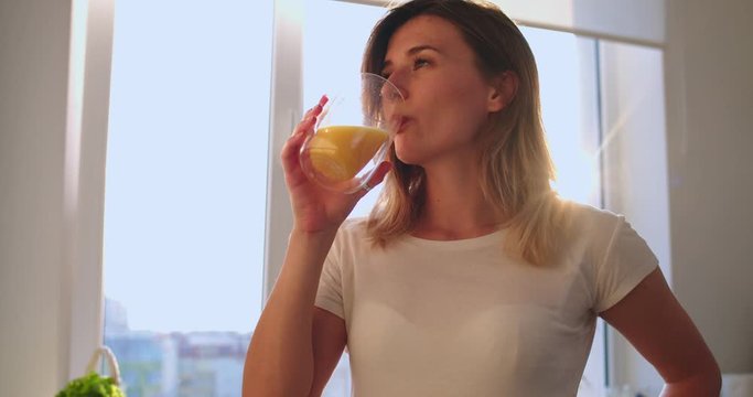 Portrait shot of the young Caucasian beauiful woman sipping an orange juice and enjoying it in the morning in the kitchen, them=n smiling to the camera. Close up.