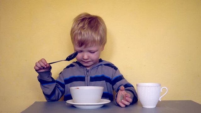 Small blond child sits at table with two cups on yellow background. Boy pours hot tea from one cup into another with spoon. Baby comes up with entertainment and is actively learning necessary skills