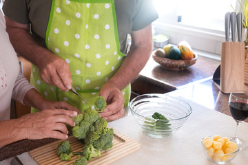 The woman's hands guide her husband's hands to cut and clean the broccoli. Stay together in the kitchen and prefer healthy vegetables