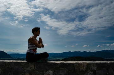 Naklejka na ściany i meble Woman sitting on a stone wall doing yoga with sky and clouds in background.
