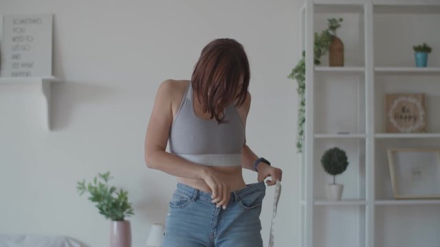 Young woman admiring the result of weight loss while wearing old jeans in bedroom. Happy female taking picture of her belly after you lost weight. 