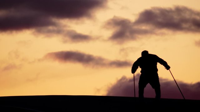 Man ride inline skates over top of hill, silhouetted telephoto shot against sunset sky. Sportsmen train at summer season, move uphill by skating and push poles