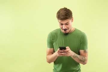 Caucasian young man's half-length portrait on green studio background. Beautiful male model in shirt. Concept of human emotions, facial expression, sales, ad. Betting, online purchases, web surfing.