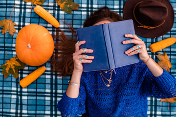 Autumn woman in knitted sweater enjoes of reading book on a blue cozy plaid blanket covered with dry maple leaves. Autumn time. Top view