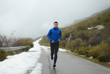 Athlete running under the snow on winter mountain road. Cold season outdoor training.