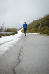 Athlete running under the snow on winter mountain road. Cold season outdoor training.