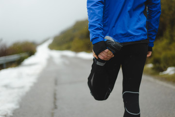 Detail of athlete warming up and stretching before running under the snow in winter mountain road.