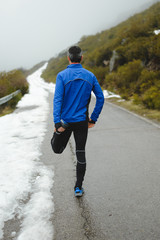 Back view of athlete warming up and stretching legs before running under the snow in winter mountain road.