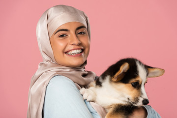 happy arabic young woman in hijab holding cute corgi puppy, isolated on pink