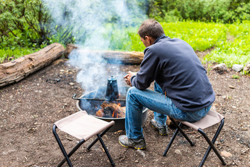 Campsite with man sitting on chair warming by firepit smoke and teapot on grill in Albion Basin, Utah summer in Wasatch mountains in evening