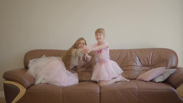 Young caucasian mother and her cute little girl in the same skirts jumping on the leather sofa and turning on TV. Woman and her daughter having fun together indoors