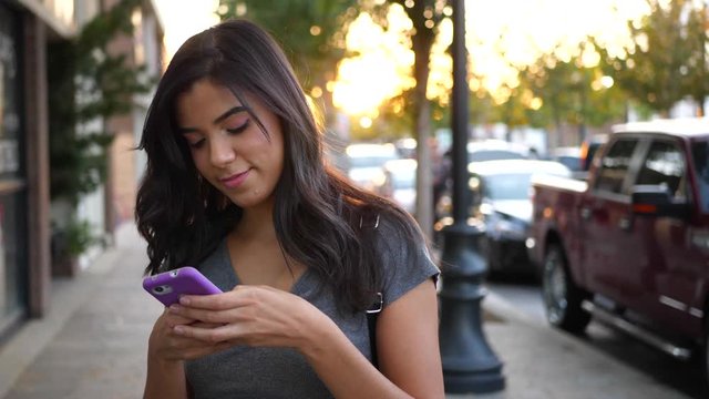 A hispanic woman walking on urban city streets texting and looking happy at a message on her cell phone SLOW MOTION.
