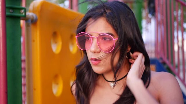 A young hispanic woman hipster wearing vintage fashion clothing and retro pink aviator sunglasses in a park playground SLOW MOTION.