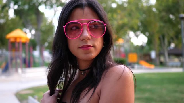 A young woman hipster wearing vintage fashion clothing and retro pink aviator sunglasses in a park playground SLOW MOTION.