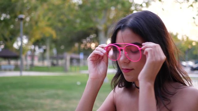 A young hispanic woman hipster wearing vintage fashion clothing and retro pink aviator sunglasses in a park playground SLOW MOTION.