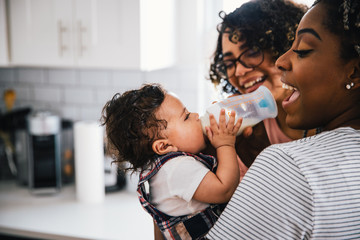 Playful loving mother feeding milk to cute son by girlfriend in kitchen