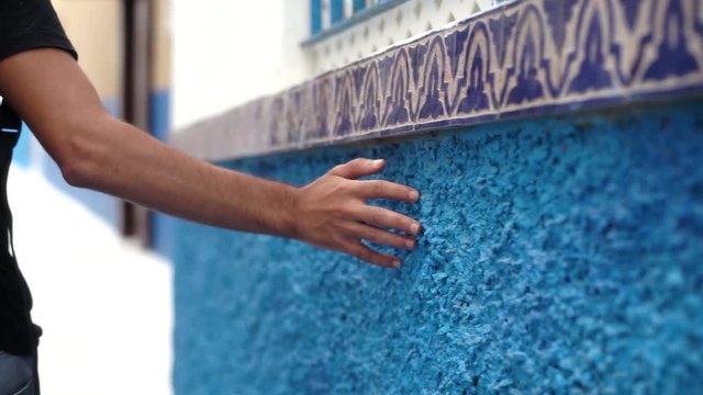 man walks and grazes hand on blue wall in Chefchaouen, Morocco.
