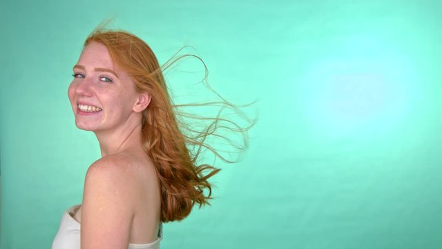 Smilling red-haired woman is standing in front of a green screen while wind blows up her long hair.