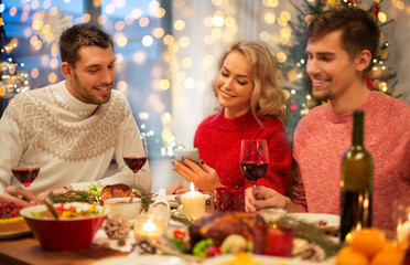 holidays and celebration concept - happy friends with smartphone having christmas dinner at home