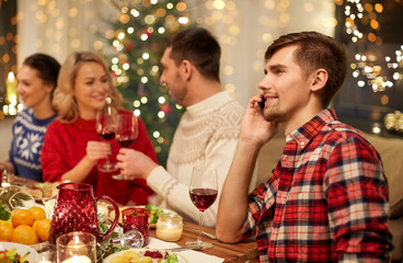 holidays, communication and celebration concept - happy young man calling on smartphone and having christmas dinner with friends at home