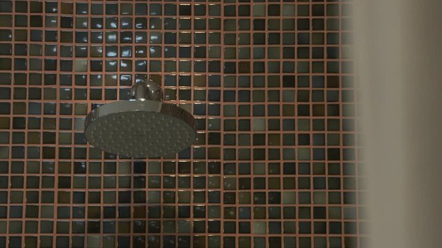 An extreme closeup shot with a racking focus of a white bath towel and a stainless steel shower head mounted on a wall covered with mosaic tiles inside the bathroom of a luxurious hotel..