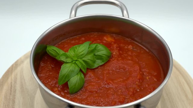 Boiled homemade tomato sauce in cooking pan with green basil leaves. Hot tomato sauce rotating on white background. Fresh cherry tomatoes, garlic and extra vigrin olive oil. Italian cuisine concept
