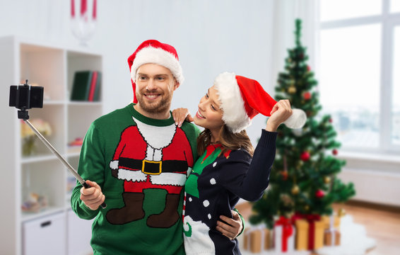 celebration, technology and winter holidays concept - happy couple in santa hats taking picture by smartphone on selfie stick at ugly sweater party over christmas tree at home background