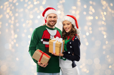 people and holidays concept - happy couple in santa hats with christmas gifts at ugly sweater party over festive lights background