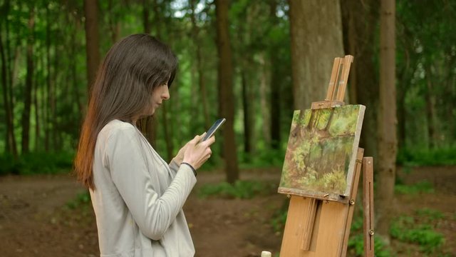The girl artist completed the work on her painting and chatting in her smartphone. A beautiful brunette woman is chatting with her friends on a social network about her hobby - painting in nature.