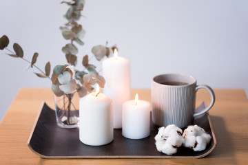 decoration, hygge and cosiness concept - burning white candles, tea in mug, branches of eucalyptus populus and cotton flowers on table
