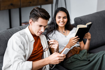 multicultural students holding book and using smartphone in apartment