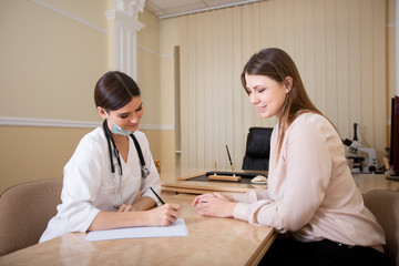 Female doctor sitting at the desk in the office and writing a prescription to the patient.