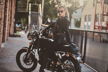 Fototapeta na wymiar Stylish female biker is sitting at her black bike while posing for a photoshoot on the street.