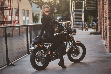 Fototapeta na wymiar Stylish female biker is sitting at her black bike while posing for a photoshoot on the street.