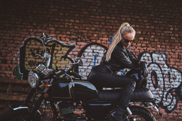 Fototapeta na wymiar Stylish sexy woman in biker clothing is posing for photographer next to her bike and graffiti wall.
