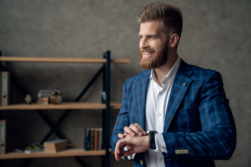 Portrait of smart responsible man with modern hairdo looking at watch on wrist over blurred office...