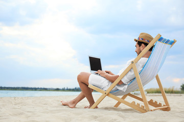Young man with laptop sitting in deck chair on sea beach. Space for text