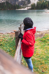 Women with red rain jacket standing all alone on a wooden fence at a mountain lake bank on a cloudy misty rainy autumn day and enjoying the peacful nature view after a hike.