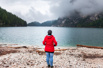 Fototapeta na wymiar Women with red rain jacket standing all alone on a rocky mountain lake bank on a cloudy misty rainy autumn day and enjoying the peacful nature view after a hike.