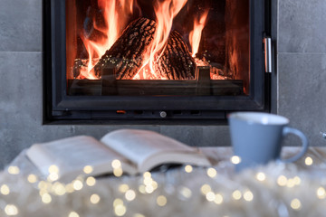 Hygge concept with open book and cup of tea near burning fireplace