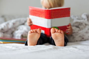 Sweet toddler boy, reading book at home, sitting in bed, lots of books