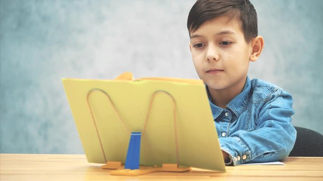 Young concentrated school kid in blue jeans t-shirt writing, copying the text from book on the bookholder.