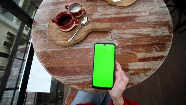 Top view of woman hands holding smartphone mobile with green screen spending time in modern cafe coffee shop outdoors on the roof. Chroma-key touchscreen, mock-up, free education application.