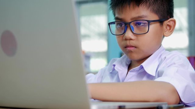 A boy who is Asian elementary school student in a white school uniform and wearing glasses, is using a laptop, searching for knowledge on the internet.
