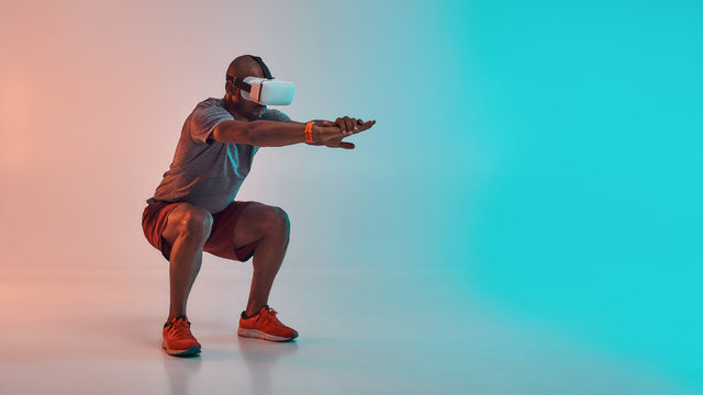 Full length of young african man in sports clothing doing squat while wearing virtual reality glasses
