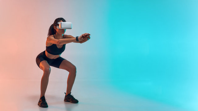 Full length of young sporty woman in sports clothing doing squat while wearing virtual reality glasses