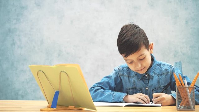 Young concentrated school kid in blue jeans t-shirt writing, copying the text from book on the bookholder. His sister joining him, looking how he is writing. He fends her off.